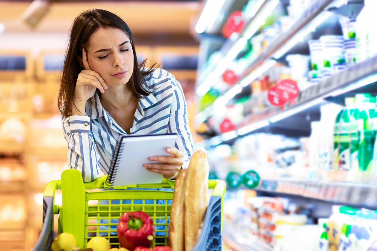 Una donna osserva attentamente la sua lista della spesa mentre si trova in un supermercato, con un carrello pieno di frutta, verdura e pane. Sembra concentrata, probabilmente pianificando i suoi acquisti per rispettare il budget
